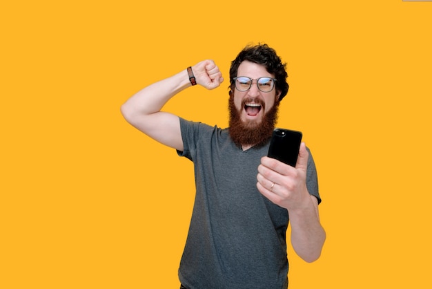 Фотография бородатого парня, держащего mobioe и празднующего с поднятой рукой на желтом