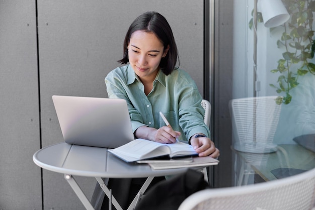 Photo of beautiful asian student watches training webinar on\
laptop computer writes down important information in notebook poses\
at table in cozy room female copywriter watchs website video