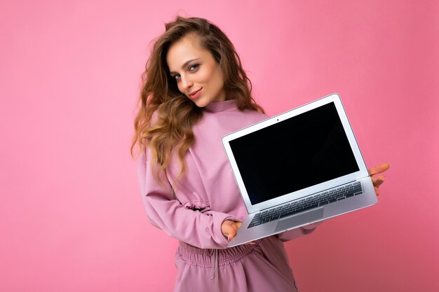 Photo of beautiful blond curly young woman lookind at camera holding computer laptop with empty