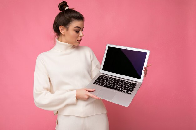 Photo of beautiful brunet young woman with gathered dark hair wearing white sweater holding computer laptop and looking at the netbook isolated on pink wall background.