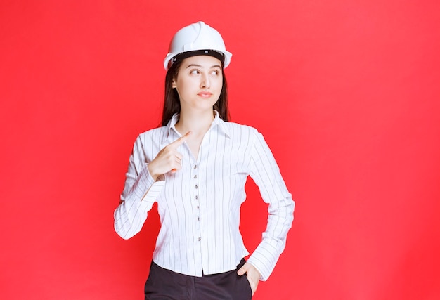 Photo of a beautiful business woman wearing safety hat pointing away with fingers.