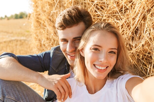 Foto di bella coppia uomo e donna prendendo selfie mentre era seduto sotto un grande pagliaio in campo dorato, durante la giornata di sole