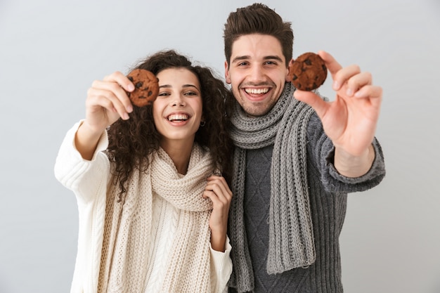 灰色の壁に隔離された、クッキーを保持しているスカーフを身に着けている美しいカップルの男性と女性の写真