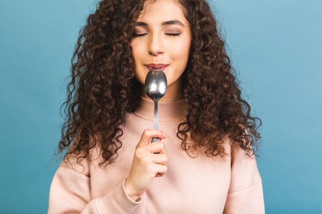 口の中で金属のスプーンを保持している美しい巻き毛の女性の写真は、おいしい食べ物の孤立した青い背景を夢見て空のスペースを検索します。