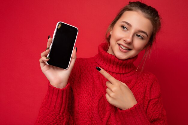 赤い背景の上に分離された暖かい赤いセーターを着て美しいかわいい笑顔の若い女性の写真