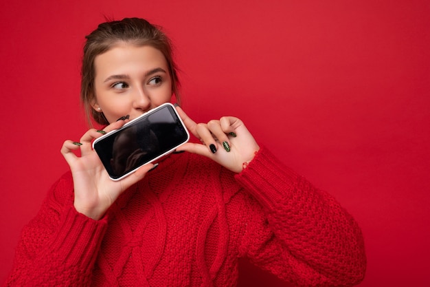 빨간 배경 벽에 격리된 따뜻한 빨간 스웨터를 입은 아름다운 젊은 여성의 사진은 스마트폰을 들고 옆을 바라보는 흉내내기용 빈 디스플레이가 있는 전화기를 보여줍니다.