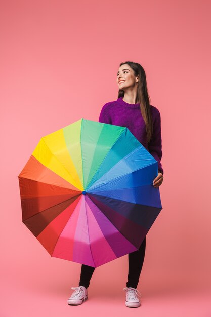 Photo of a beautiful emotional young woman posing isolated over pink space holding rainbow umbrella.