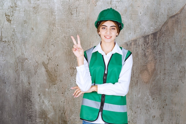 Photo of beautiful female architect in green helmet showing victory sign