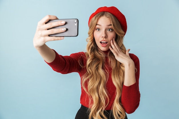 Photo of beautiful girl 20s wearing red beret taking selfie on mobile phone isolated 