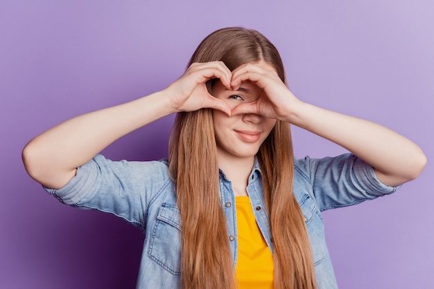 Photo of beautiful girl making heart gesture cover eye on purple background