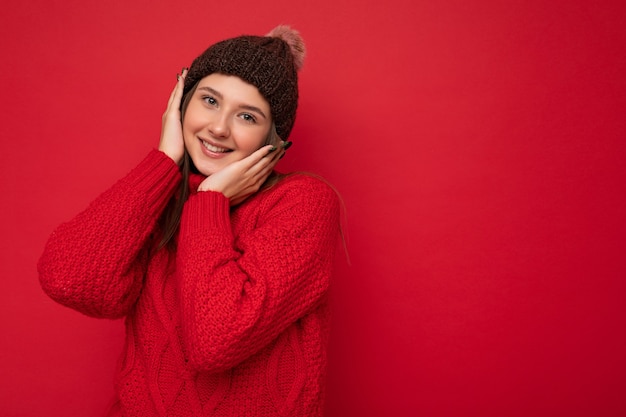 赤いセーターと帽子をかぶってカメラを見て赤い背景の壁に孤立して立っている美しい幸せな笑顔の若いブルネットの女性の写真。コピースペース