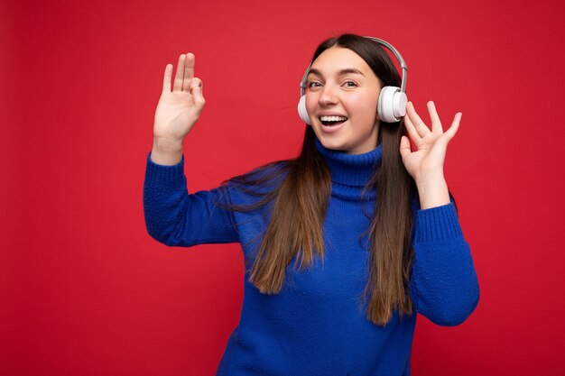 Photo of beautiful happy smiling young brunette woman wearing blue sweater isolated over red background wall wearing white bluetooth headphones listening to cool music and dancing looking at camera.