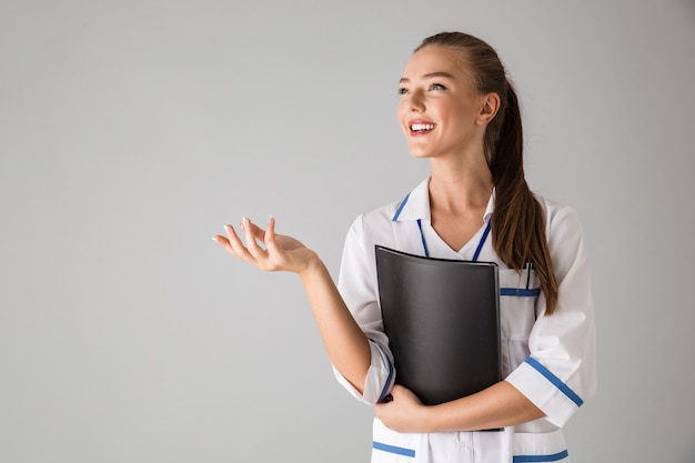 Photo of a beautiful happy young woman cosmetologist doctor isolated over grey wall  holding folder.