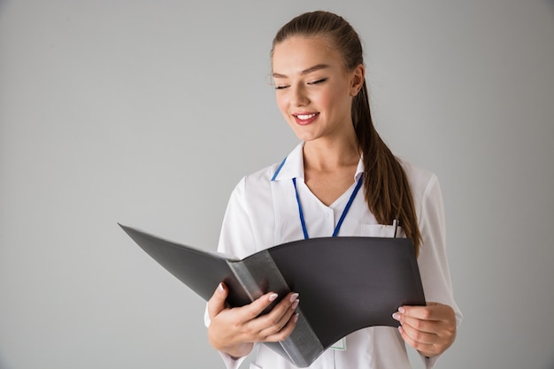 Photo of a beautiful happy young woman cosmetologist doctor isolated over grey wall  holding folder.