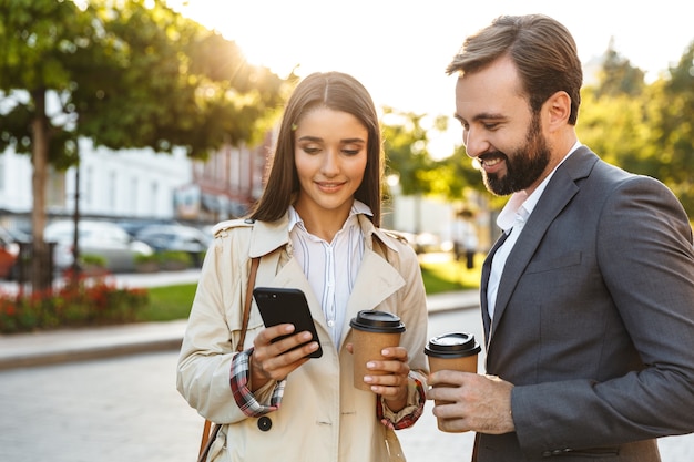 Photo of beautiful office workers man and woman in formal wear holding takeaway coffee while using mobile phone on city street