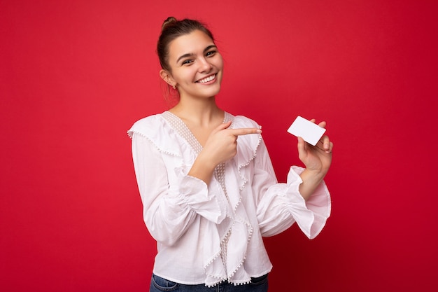 Photo of beautiful positive smiling young dark blonde woman wearing white blouse isolated over red background holding credit card looking at camera and pointing finger at plastic contactless card.