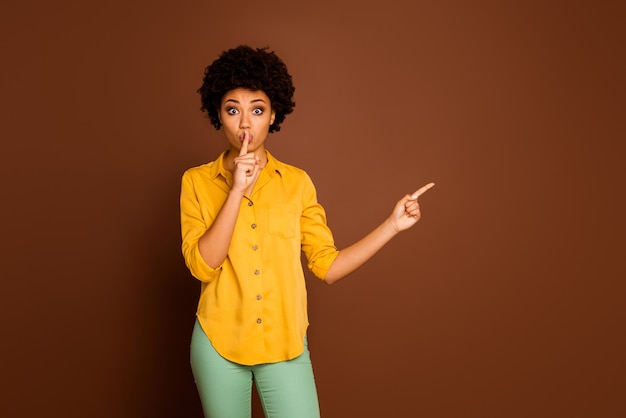 Photo of beautiful pretty dark skin lady indicating fingers empty space finger on lips advising sale shopping prices wear yellow shirt green trousers isolated brown color
