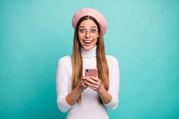 아름다운 예쁜 아가씨가 전화를 손에 들고 있는 사진은 좋은 소식을 읽는 열린 입 배너 착용 사양 현대적인 분홍색 베레모 모자 흰색 터틀넥 격리된 청록색 배경