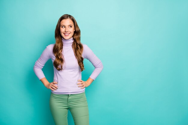 Photo of beautiful pretty lady long curly hairstyle good mood hands by sides look empty space wondered wear lilac purple sweater green trousers isolated pastel blue color