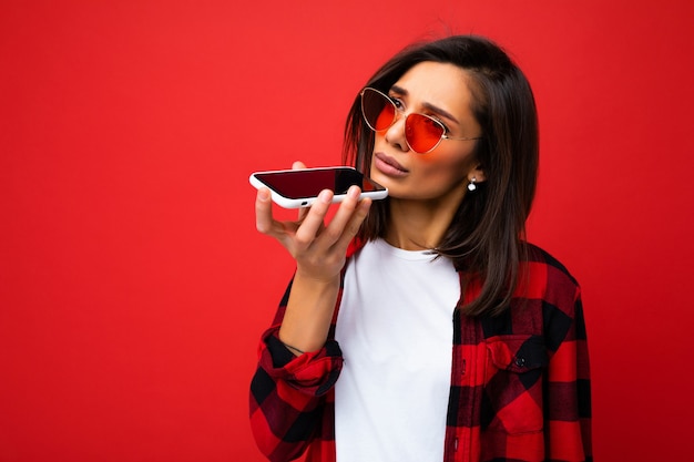 Photo of beautiful sad young brunet woman wearing stylish red shirt white t-shirt and red sunglasses isolated over red background using mobile phone recording voice message looking to the side.