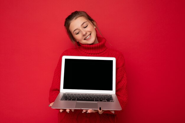 빨간 벽 배경 위에 절연 빨간 스웨터를 입고 netbook 화면에서 아래를 내려다 보면서 컴퓨터 노트북을 들고 아름 다운 만족 된 행복 한 젊은 여자의 사진. 빈 공간