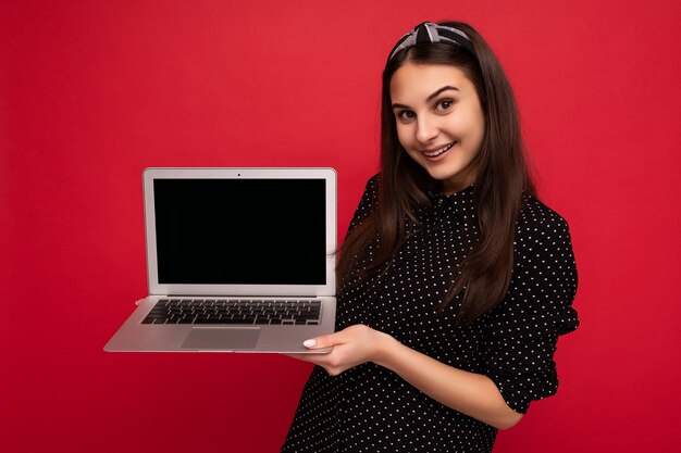 화려한 벽 배경에 격리된 카메라를 바라보는 컴퓨터 노트북을 들고 검은 옷을 입고 웃고 있는 아름다운 브루넷 소녀의 사진. 빈 공간