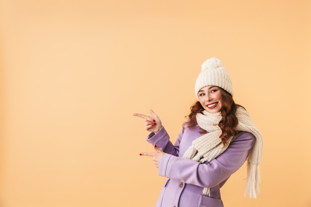 Photo of beautiful woman 20s in winter clothes pointing fingers aside, while standing isolated 