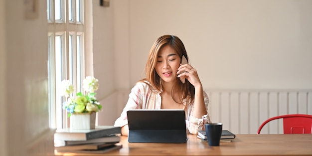 Photo a beautiful woman using a smartphone to calling someone while sitting in front a computer tablet with keyboard case at wooden working table over comfortable living room