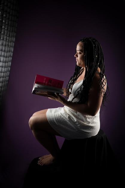 photo of beautiful woman with braids in her hair sitting wearing white clothes reading a book studio shot with dark lilac background positive person