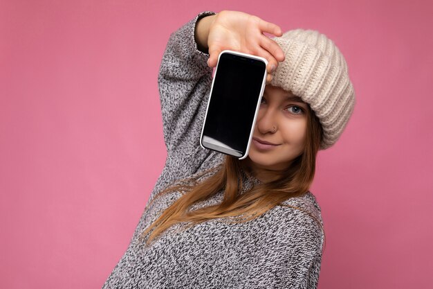 Foto di bella giovane donna bionda che indossa un maglione grigio casual e cappello beige isolato sopra il muro rosa tenendo in mano e mostrando il telefono cellulare con display vuoto.