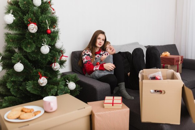 近くのクリスマスツリーと一緒に時間を過ごしながら自宅のソファに座っている美しい若いカップルの写真