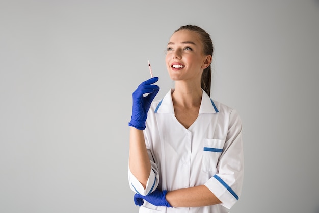 Photo of a beautiful young woman cosmetologist doctor isolated over grey wall  holding injection syringe.