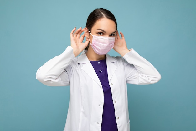 Фото красивой молодой женщины-врача в белом халате и медицинской маске, стоящей изолированной над синим
