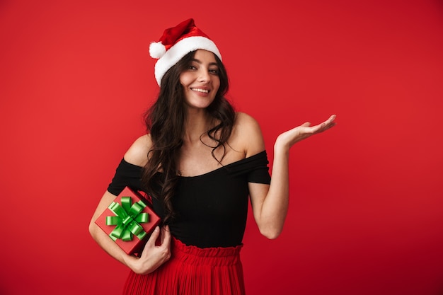 Photo of a beautiful young woman standing isolated over red wall holding gift box wearing christmas hat.