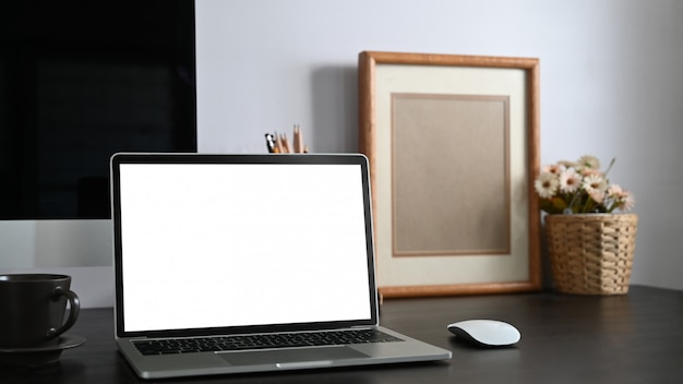白い空白の画面コンピューターラップトップ、書籍、ノートブック、鉛筆ホルダー、額縁、鉢植えの植物と一緒に黒いセメントの壁と一緒に置く黒い作業机の写真。