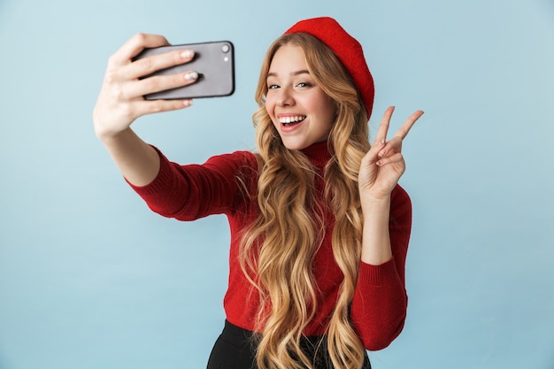 Photo of blonde girl 20s wearing red beret taking selfie on mobile phone isolated 