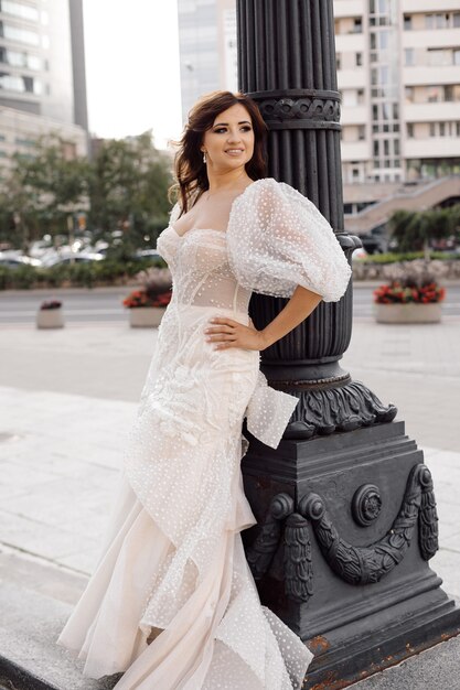 Photo of bride with dark hair in white wedding dress at lamppost in background of city buildings, looks happily. Happy moments.