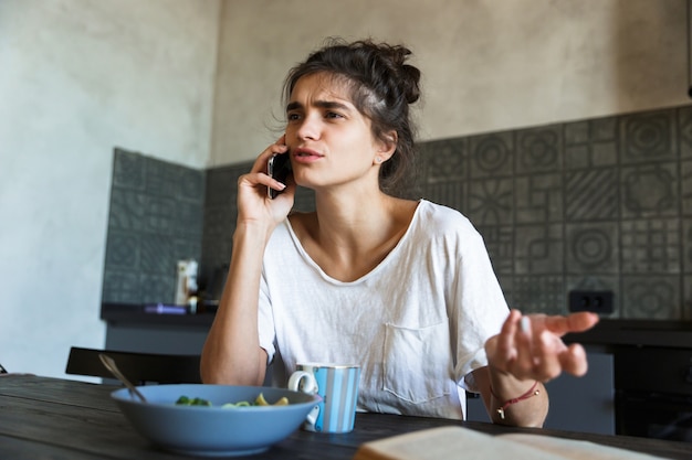 Foto di una donna bruna cova che legge un libro e parla al cellulare mentre fa colazione in cucina a casa