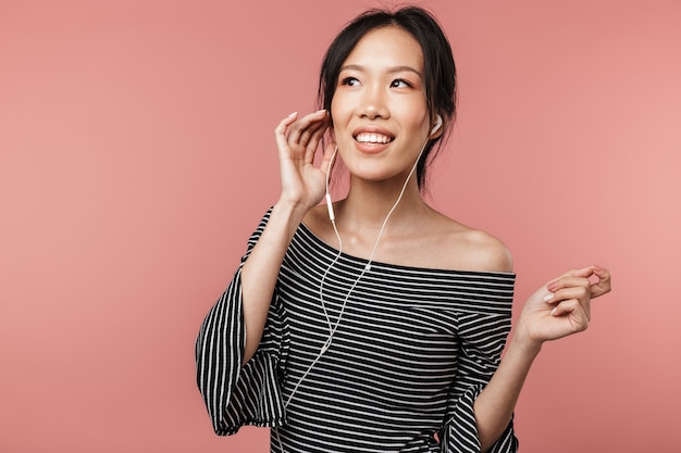 Photo of brunette asian woman dressed in basic wear singing and listening to music with earphones isolated over red wall
