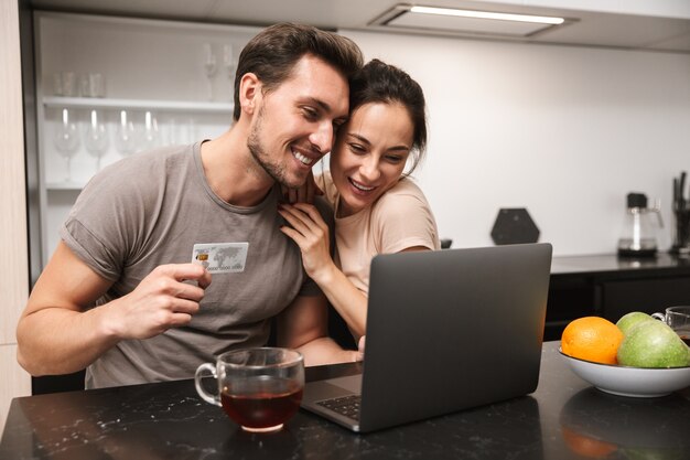 Photo of brunette couple man and woman using laptop with credit card, while sitting in kitchen