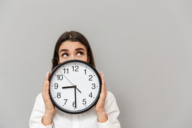 Photo of business woman in white shirt looking upward while covering face with big round clock, isolated over gray wall