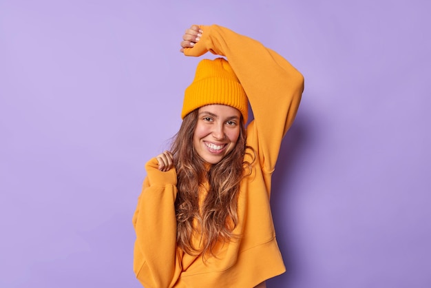 Photo of carefree woman with long hair smiles happily wears casua orange jumper and hat keeps arm raised dances with rhythm of music isolated over purple background has fun. Happiness concept