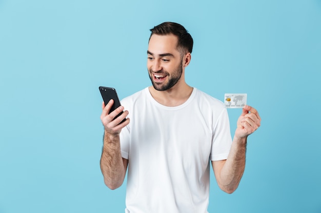 Photo of caucasian brunette man wearing basic t-shirt smiling while holding smartphone and credit card isolated over blue 