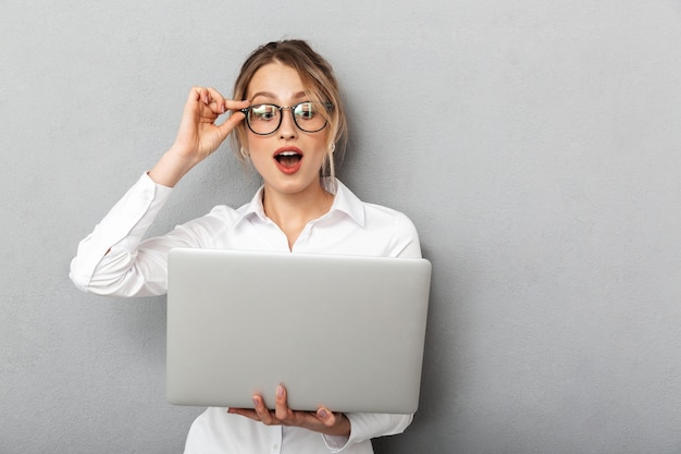 Photo of caucasian businesslike woman wearing glasses standing and holding laptop in the office, isolated 