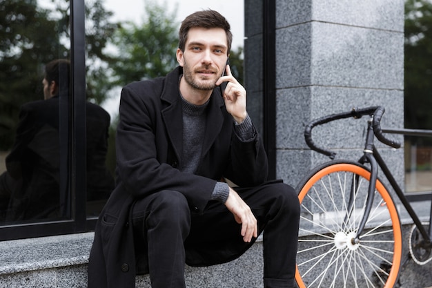 Фотография кавказца 20 лет, использующего мобильный телефон, сидя на велосипеде на улице