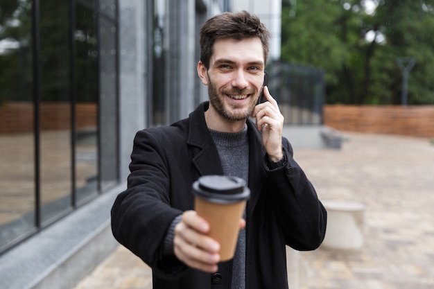 Photo of caucasian man 20s using mobile phone, while walking outdoor with takeaway coffee