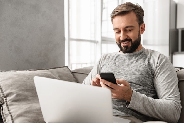 Foto dell'uomo caucasico 30s nell'abbigliamento casual facendo uso dello smartphone, mentre lavorando al computer portatile in salone