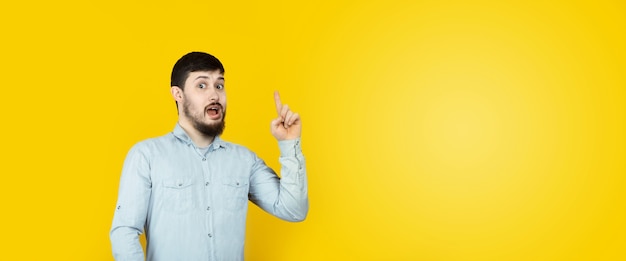 Photo of caucasian man  reliable person got new brilliant idea, startup raise finger, wear jeans denim shirt  over yellow background, panoramic mock-up