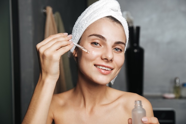 Photo of caucasian woman with towel on head applying cosmetic oil on her face