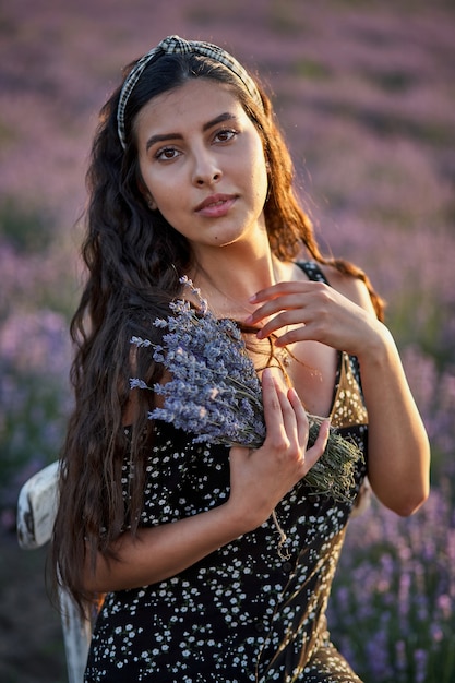 Photo of caucasian young woman in dress holding bouquet of flowers, outdoor through lavender field in summer